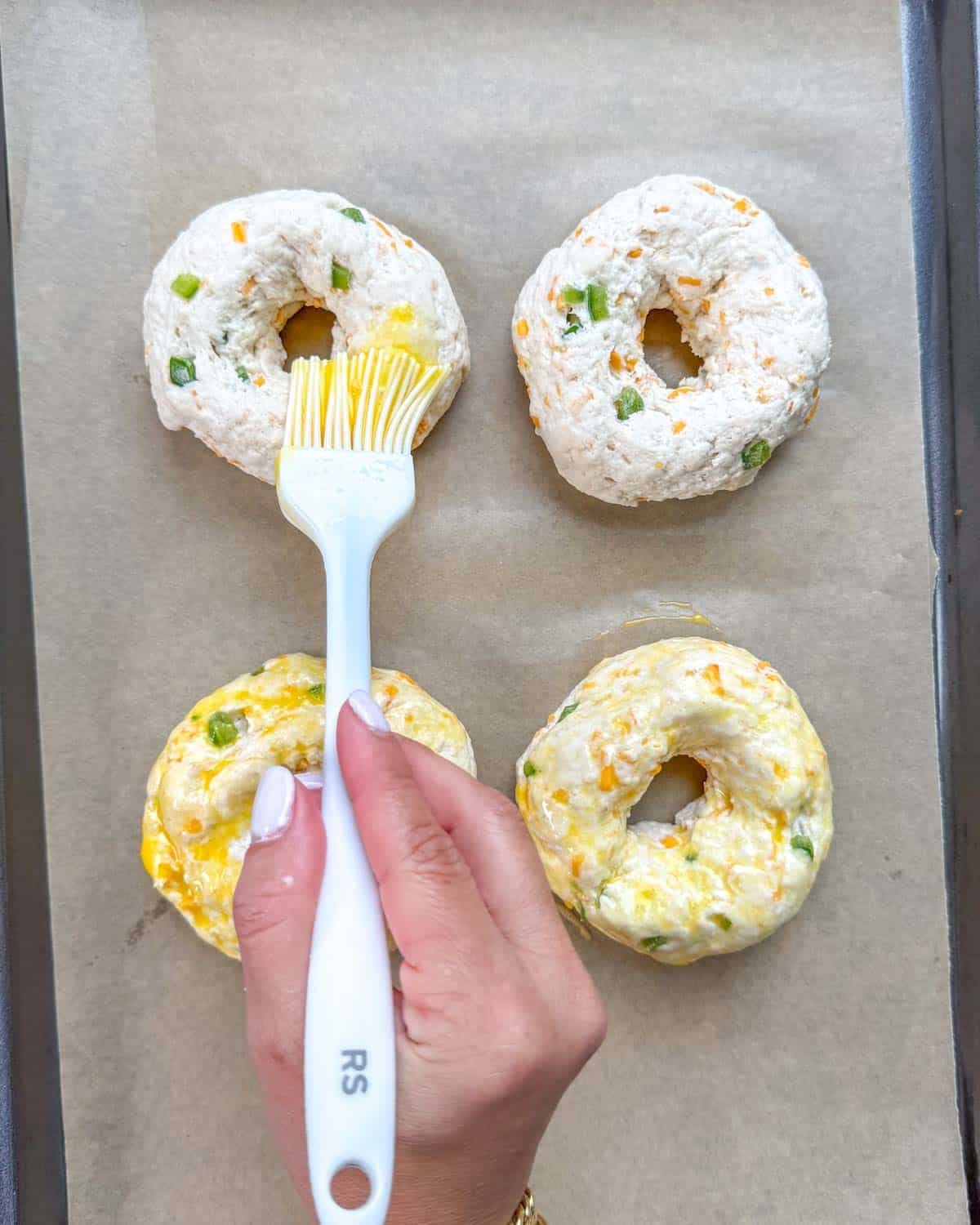 Zesty Jalapeno Cheddar Bagels 16 Egg wash being brushed onto four bagel doughs on a parchment-lined baking sheet.