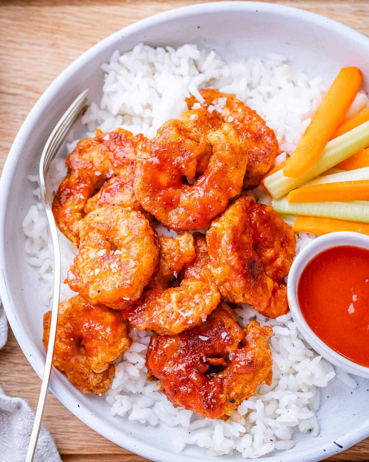 A bowl of buffalo shrimp over rice with celery, carrot and a small bowl of extra sauce