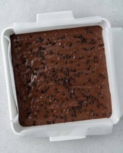 Brownie batter spread evenly in a lined square baking pan, topped with chocolate chips before baking.