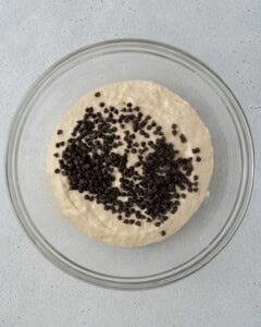Glass bowl filled with muffin batter topped with mini chocolate chips, ready to be folded in