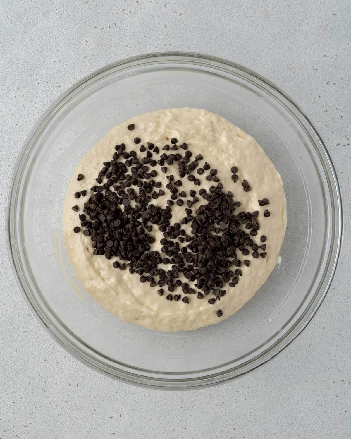 Glass bowl filled with muffin batter topped with mini chocolate chips, ready to be folded in