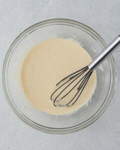 Whisking wet ingredients including yogurt, eggs, vanilla, milk, and sugar in a glass bowl.