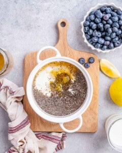 Hand pouring milk into the chia pudding mixture in a white bowl, surrounded by blueberries, honey, and lemons.