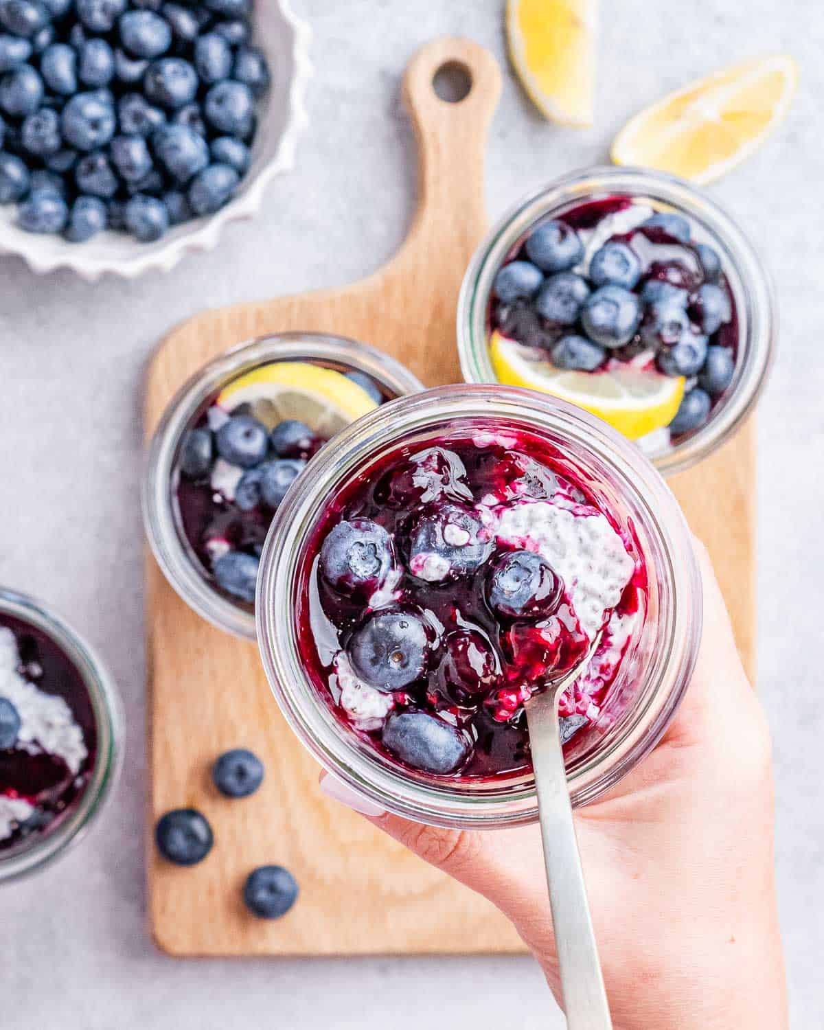 Overhead view of a hand spooning blueberry compote into a glass jar filled with chia pudding.