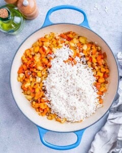 Rice added to sautéed vegetables in a skillet, ready to be stirred and toasted.