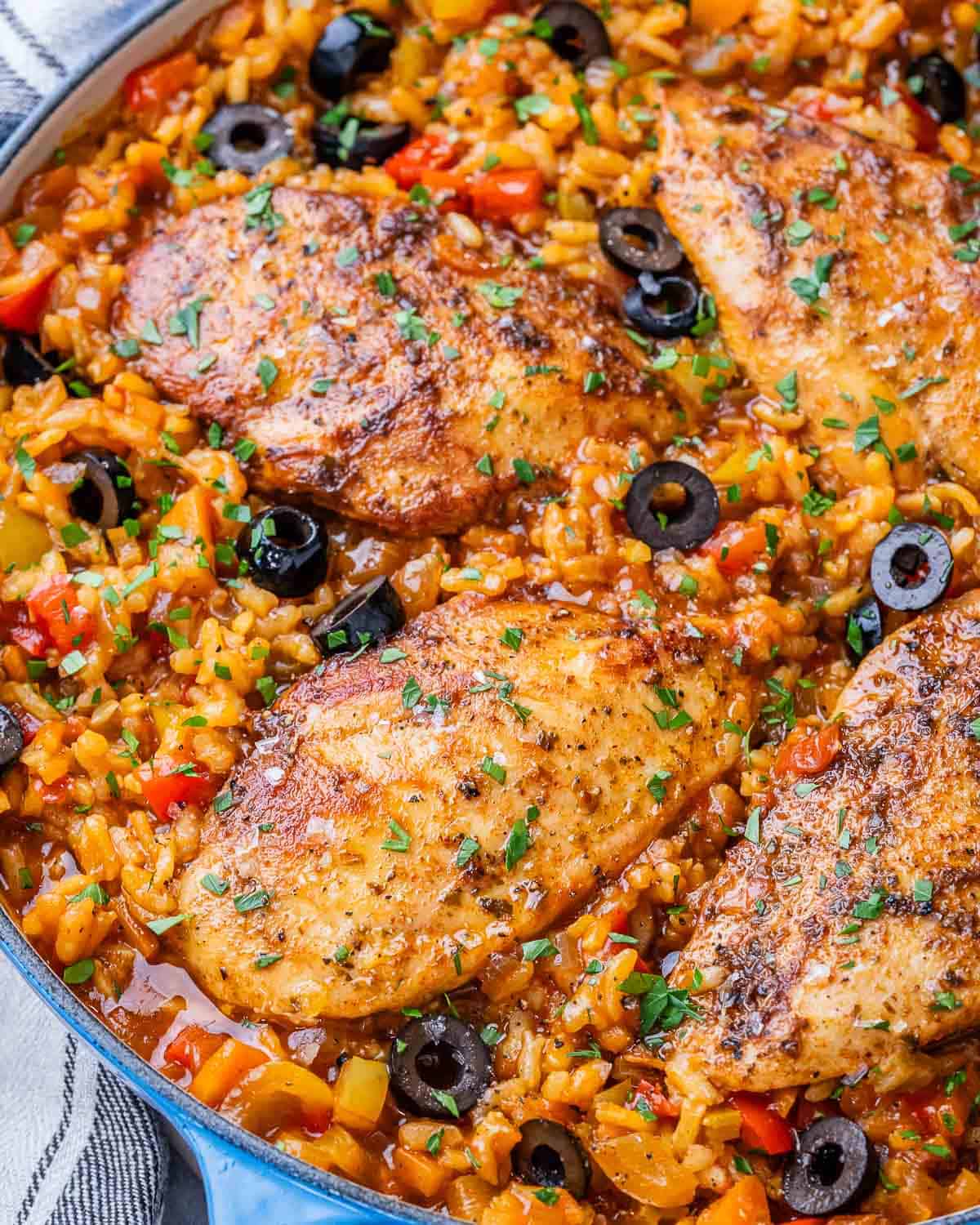 Close-up of golden, seasoned chicken breasts nestled in Spanish rice with black olives and chopped parsley.