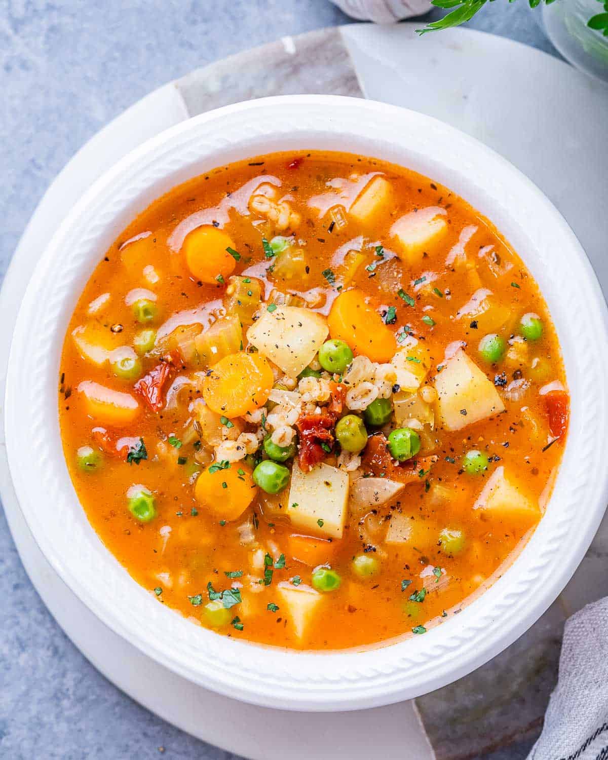 Top view of a white bowl of vegetable barley soup with barley, peas, and carrots, garnished with herbs.