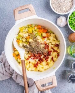 Pot with potatoes, carrots, celery, bell pepper, and dried herbs ready to be cooked into soup.