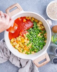 Pouring marinara sauce into a pot with peas, barley, and diced vegetables for the soup base.