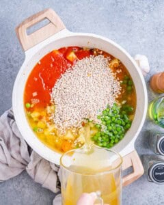 Pot filled with uncooked barley, peas, and marinara sauce being added to simmering vegetables.
