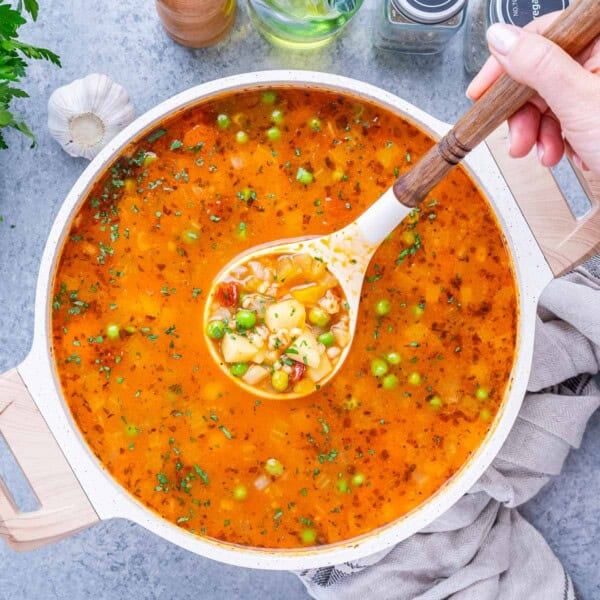 Ladle scooping vegetable barley soup from a pot, showing colorful vegetables and broth.