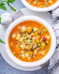 White bowl of vegetable barley soup with chunks of potato, peas, and carrots in a tomato broth.