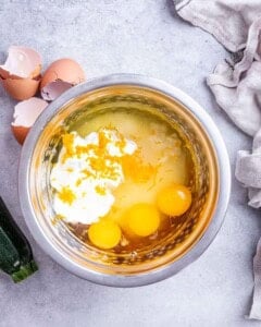 Overhead view of a mixing bowl filled with eggs, Greek yogurt, vegetable oil, and vanilla extract before whisking, with eggshells and a zucchini nearby on a light kitchen surface.