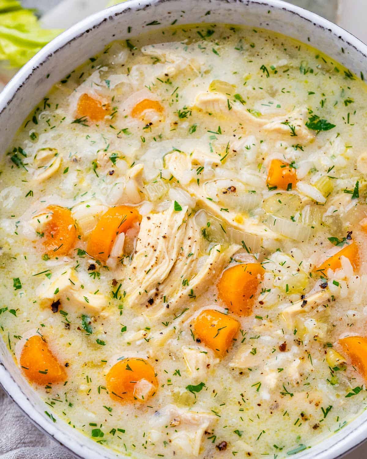 A top view close up of Avgolemono Soup showing tender chicken, orange carrot rounds, and green herbs in a lemony broth.