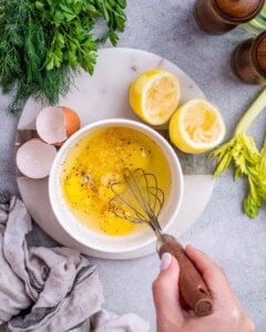 A bowl of whisked egg and lemon mixture with lemon halves, celery, and herbs nearby on the counter.