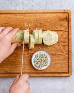 Hand slicing the chilled cookie dough log into even discs on a wooden cutting board.