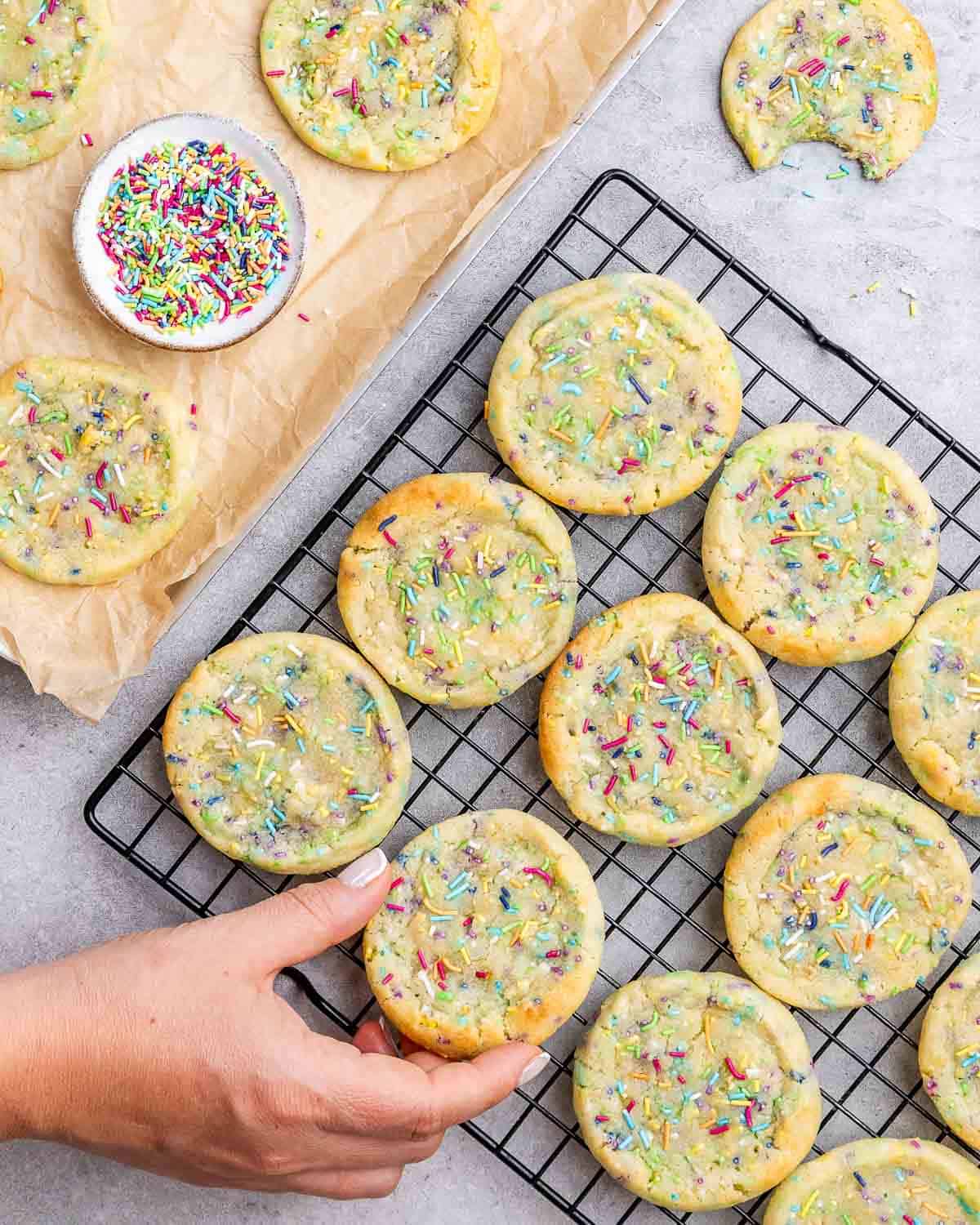 Hand placing a finished cookie onto a wire rack with a bowl of rainbow sprinkles nearby.