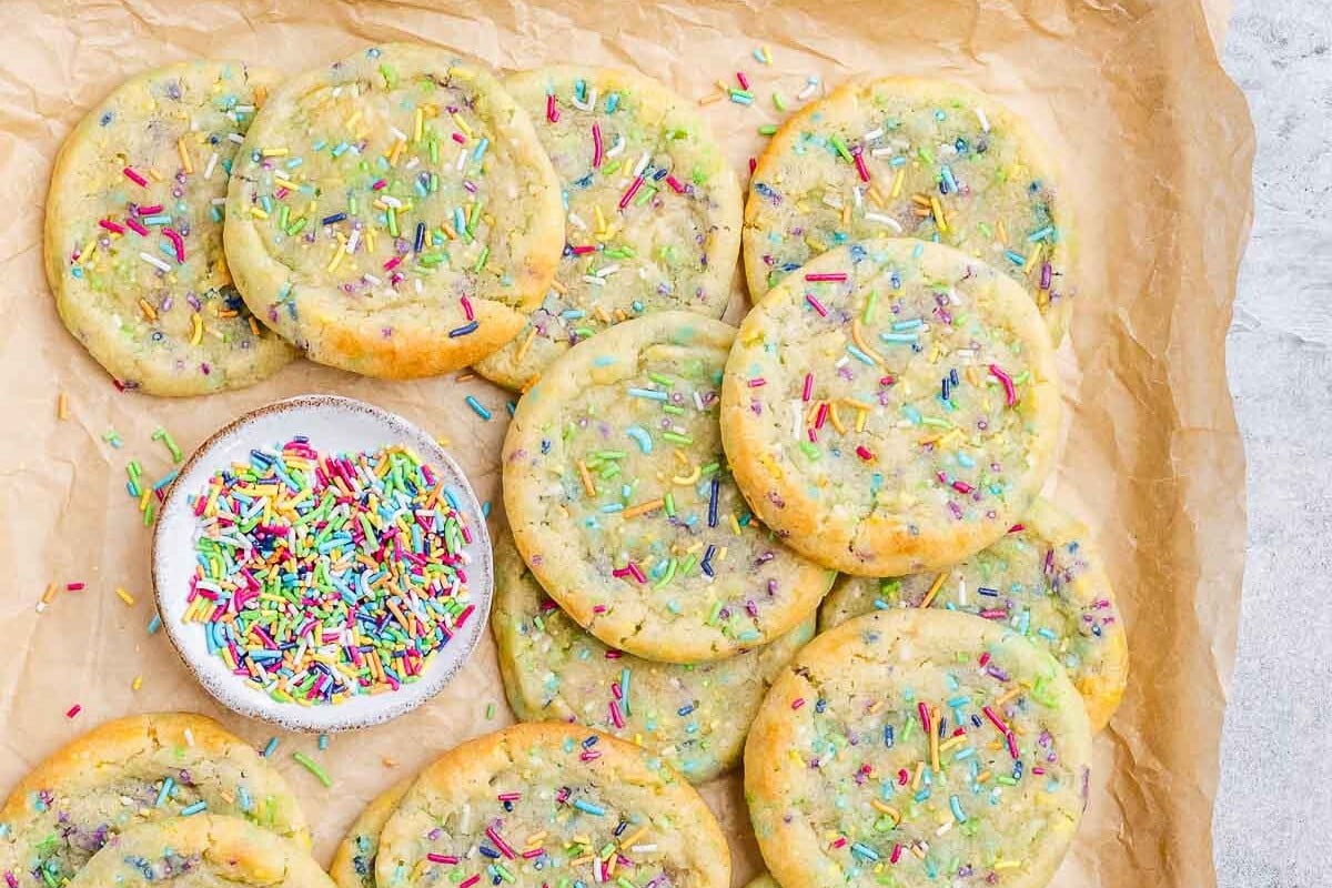 Dozens of baked birthday cake cookies on parchment paper, all topped with colorful sprinkles.