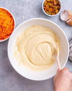 Smooth carrot cake batter in a bowl, ready to be combined with dry ingredients, with carrots and raisins in the background.