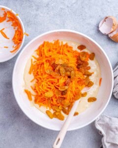 Mixing bowl with grated carrots and golden raisins being folded into batter with a spoon.