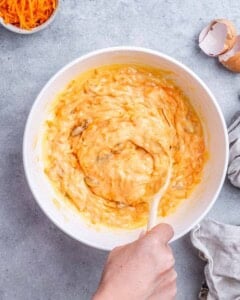 Mixing bowl with grated carrots and golden being stirred together with a wooden spoon.