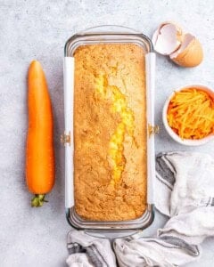 Freshly baked carrot cake loaf resting in a loaf pan, with a whole carrot and grated carrots beside it.