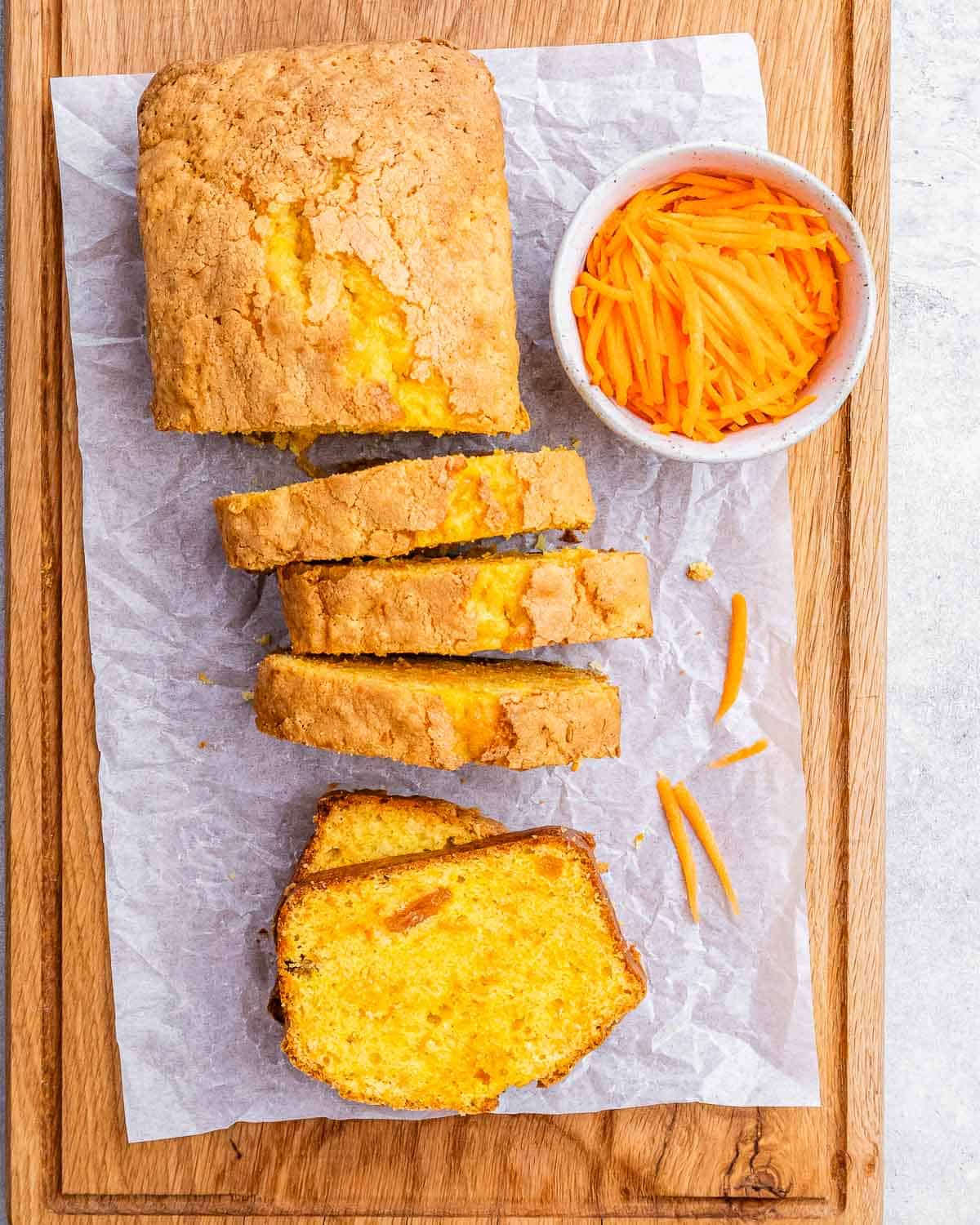 Carrot cake loaf on parchment paper, sliced into thick pieces, with a bowl of shredded carrots nearby.