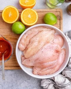 Plain raw chicken breasts in a white bowl, placed next to halved oranges and limes on a wooden board.