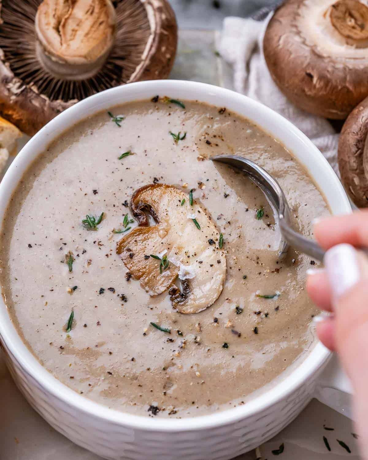 A white bowl of cream of mushroom soup with fresh mushrooms next to it and a spoon going into the soup.