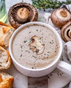 A white mug of cream of mushroom soup next to chunks of baguette and fresh mushrooms