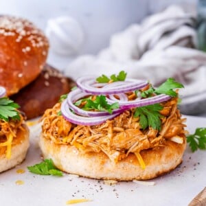 Open-faced bun loaded with BBQ shredded chicken and sliced red onion, with another sandwich in the background.