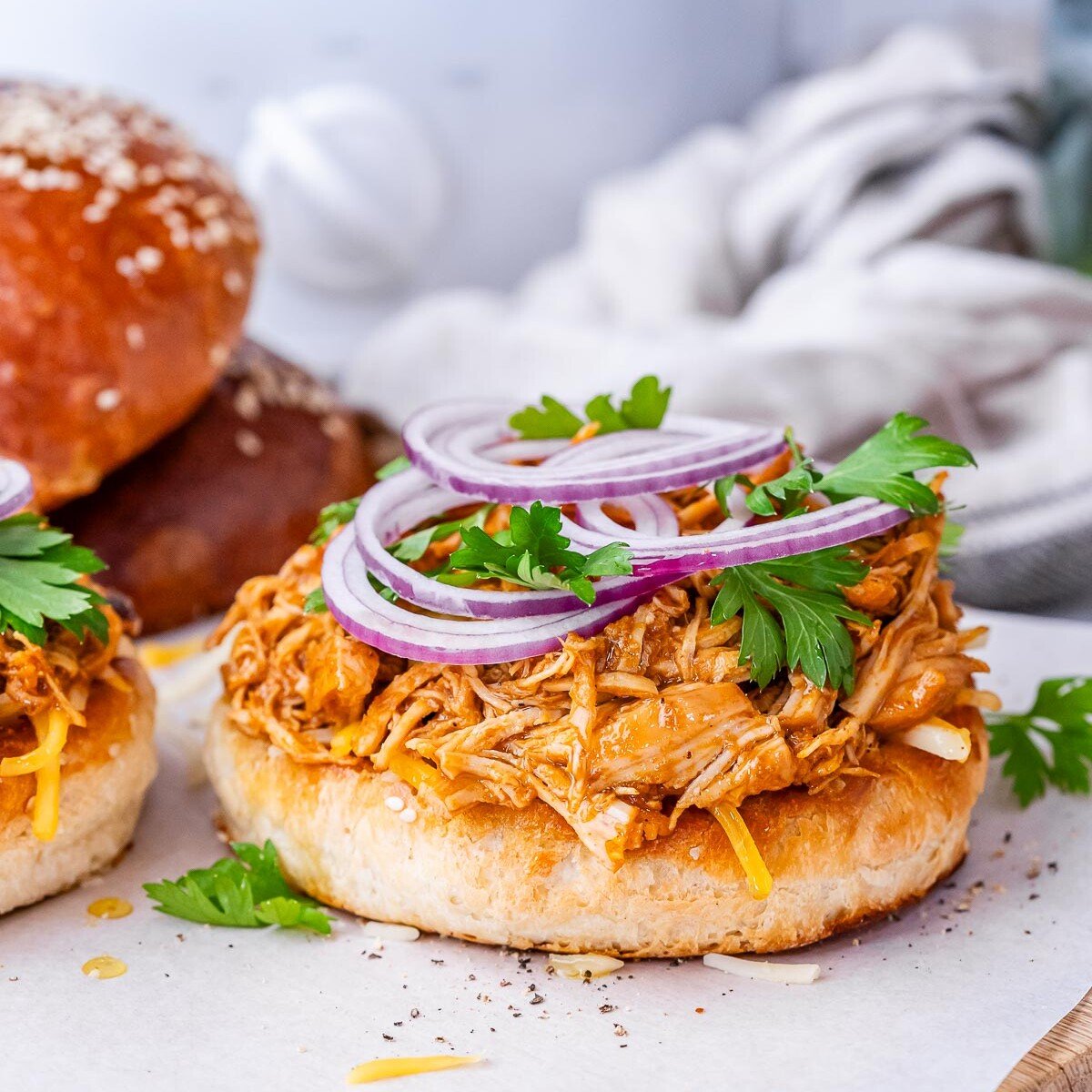 Open-faced bun loaded with BBQ shredded chicken and sliced red onion, with another sandwich in the background.