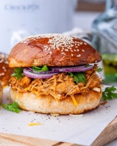 BBQ shredded chicken piled onto a sandwich bun with sesame seeds, topped with red onion and parsley.