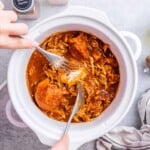Hand shredding cooked BBQ chicken with two forks inside a white crockpot.