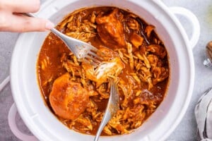 Hand using two forks to shred BBQ chicken inside a crockpot with sauce.