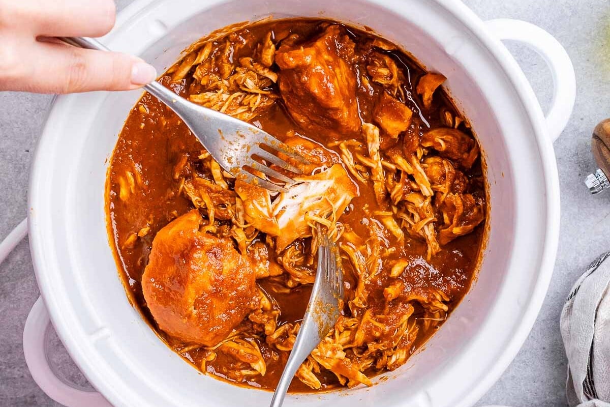 Hand using two forks to shred BBQ chicken inside a crockpot with sauce.