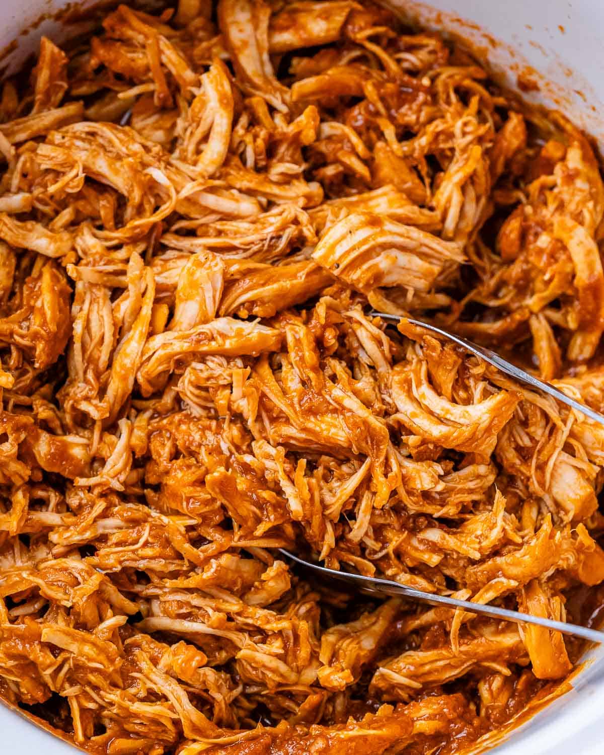 Juicy shredded BBQ chicken inside a crockpot, with metal tongs in it.