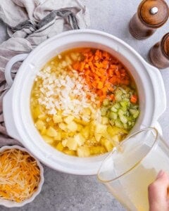 The full crockpot filled with potatoes, chopped carrots, celery, onion, with broth being poured in.