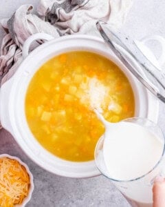 Milk being poured into the crockpot filled with broth, potatoes, carrots, and celery.