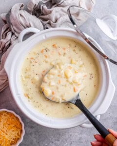 Crockpot filled with cooked diced potatoes, carrots, and broth, with a ladle lifting a creamy portion.