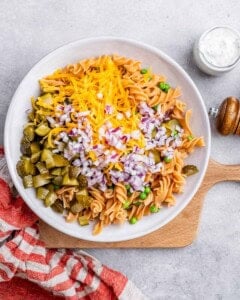 Pasta salad base in a white bowl topped with shredded cheddar, chopped red onion, and diced dill pickles before mixing.
