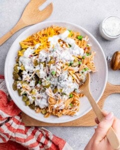 Hand stirring in creamy dressing to the pasta salad mixture in a large white bowl on a wooden board.