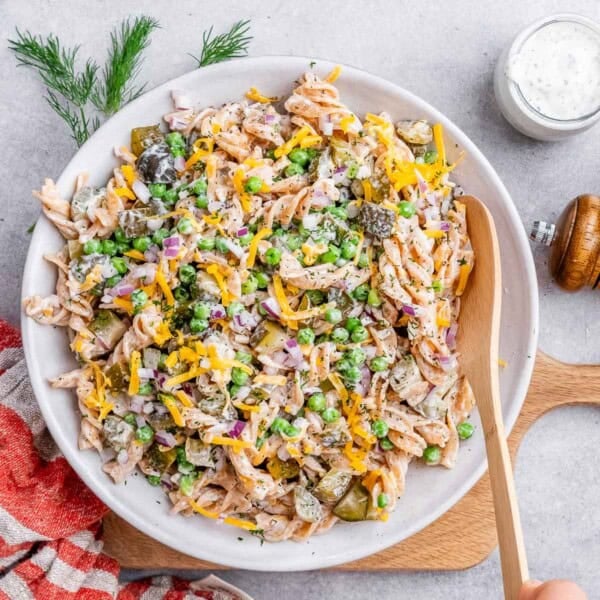 Wooden spoon stirring a large white bowl of dill pickle pasta salad, showing a well-mixed creamy texture.