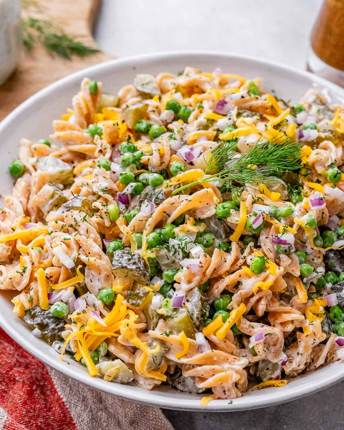 Bowl of finished dill pickle pasta salad, garnished with extra dill and peas, with a spoon ready to serve.