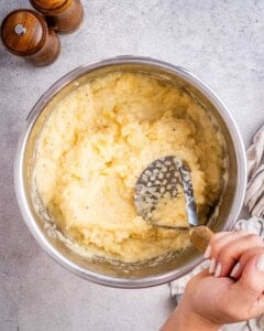 Pot of freshly mashed Yukon gold potatoes being worked with a potato masher.