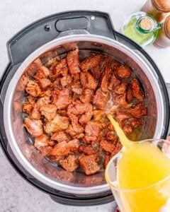 Pouring beef broth into the Instant Pot over seasoned beef chunks.