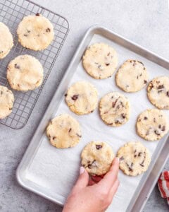 Freshly baked cookies with strawberry pieces on a parchment-lined baking sheet, being picked up by a hand.