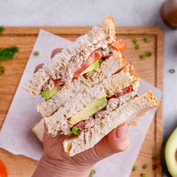 A hand holding a cut sandwich with avocado and tomato layers, placed on parchment over a wooden board.