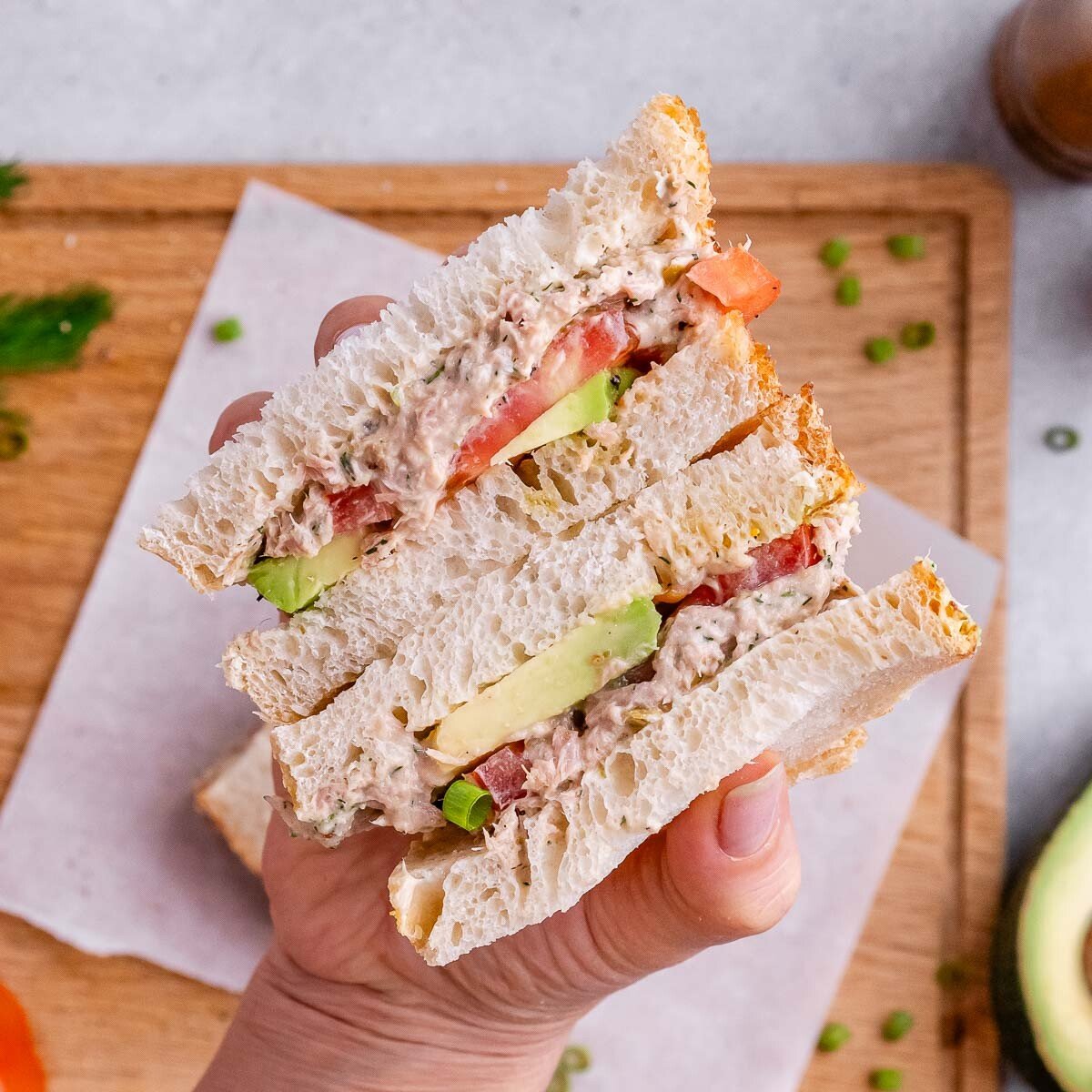 A hand holding a cut sandwich with avocado and tomato layers, placed on parchment over a wooden board.