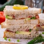Two stacked halves of a tunacado sandwich on toasted bread, garnished with a lemon slice, fresh dill, and green onion.